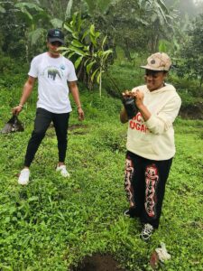 A woman plants a small tree in a green field as a man looks on.